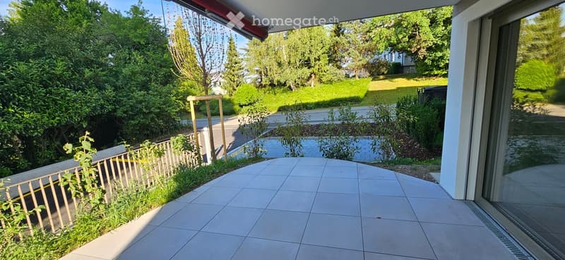Balcony area with tiled flooring, wooden railing, and surrounding greenery, including bushes and trees.