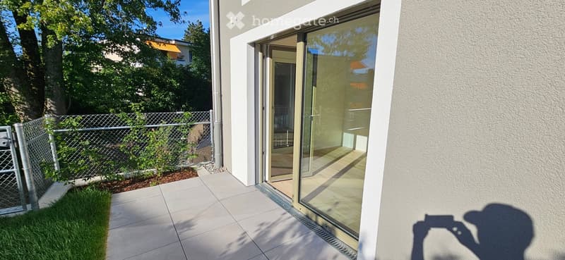 Modern balcony with large glass sliding doors, tiled floor, and a small fenced garden area with grass and plants.