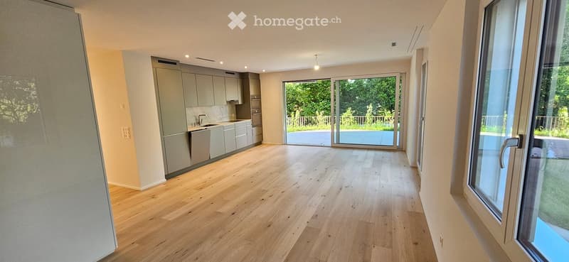Open-plan kitchen and living area with light wooden flooring, modern grey cabinets, and large glass sliding doors leading to a garden view.