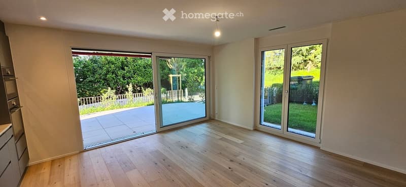 Bright, empty room with wooden flooring, large sliding glass doors leading to a patio and garden, and recessed ceiling lighting.