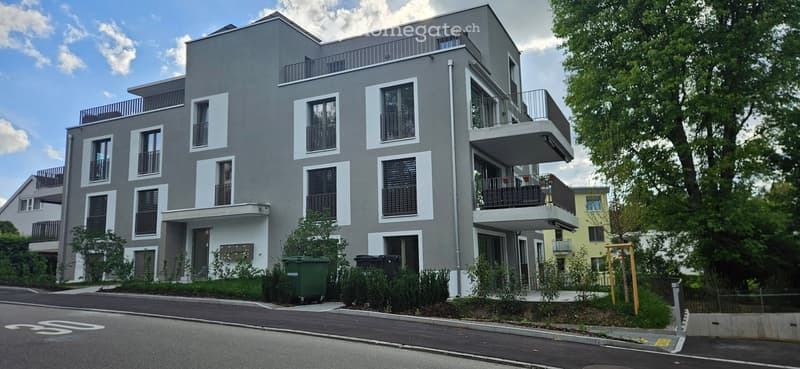 Modern multi-story apartment building with grey facade, white trim around windows, and multiple balconies. Green trash bins are placed near the entrance.
