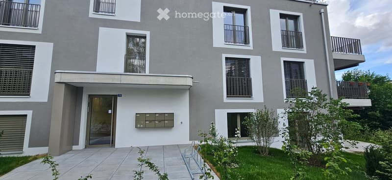 Modern multi-story building with grey facade, white trim, multiple balconies, and a small landscaped garden area in front.