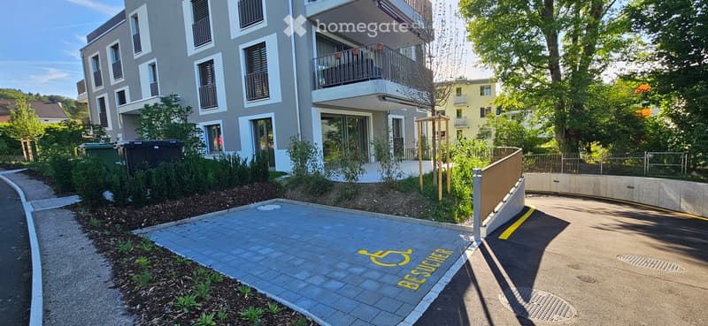 Modern multi-story residential building with balconies, surrounded by greenery and a paved parking area.