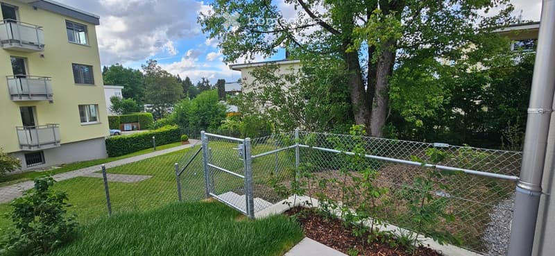 A fenced garden area with a well-maintained lawn, small plants, and a pathway leading to a multi-story residential building with balconies.