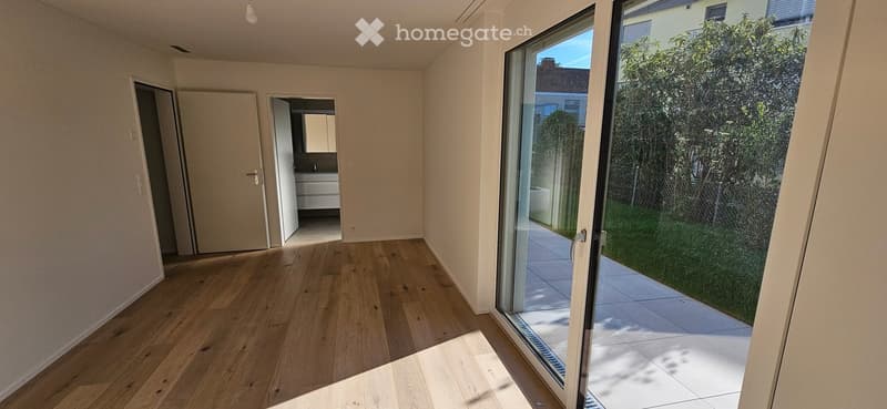 Empty room with wooden flooring, large sliding glass door leading to a patio, and visible adjacent bathroom with white cabinets and sink.