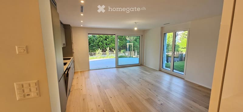 Bright, empty room with light wooden flooring, large sliding glass doors leading to a garden, and modern kitchen cabinets on one side.