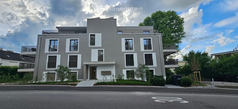 Modern multi-story residential building with grey and white facade, large windows, balconies, and surrounding greenery.