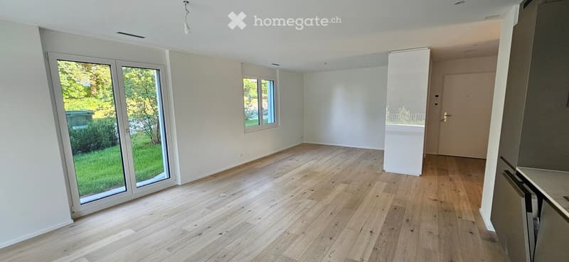 Empty room with light wooden flooring, white walls, large sliding glass door leading to a green outdoor area, and a small window on the opposite wall.