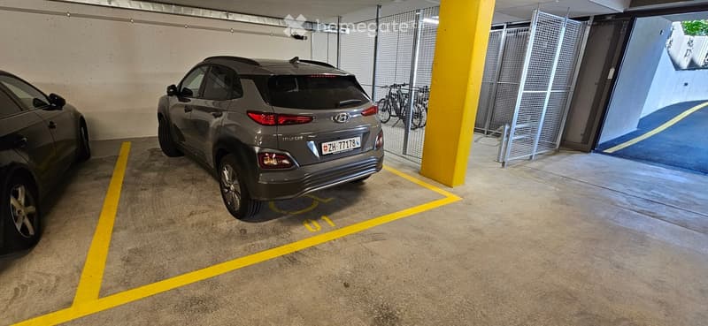 Underground parking area with marked parking spaces, a grey SUV, and bicycles stored behind a metal fence.