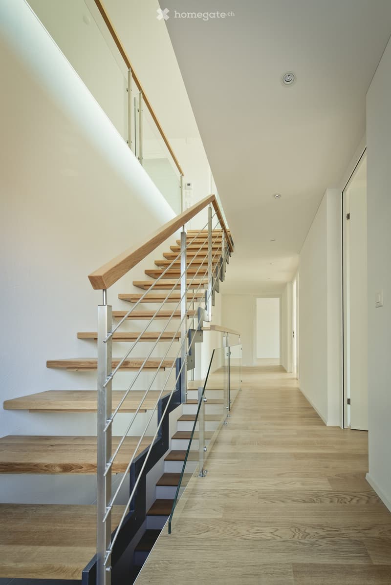 Modern hallway with wooden flooring, white walls, and a staircase featuring wooden steps and a glass railing.