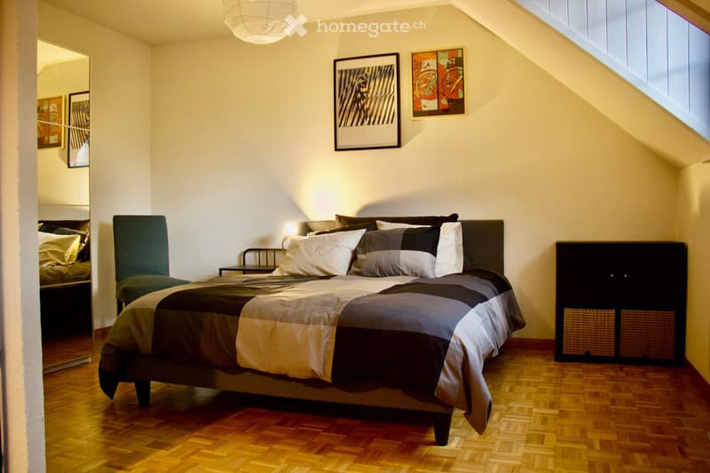 Cozy bedroom with a large bed, nightstand, chair, and cabinet. The room features wooden flooring, white walls, and modern decor including artwork and a mirror.