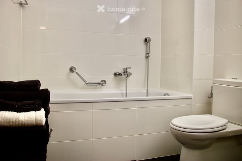 Modern bathroom with white tiled walls and floor, featuring a bathtub with a handheld showerhead, a toilet, a towel rack with towels, and a grab bar for safety.
