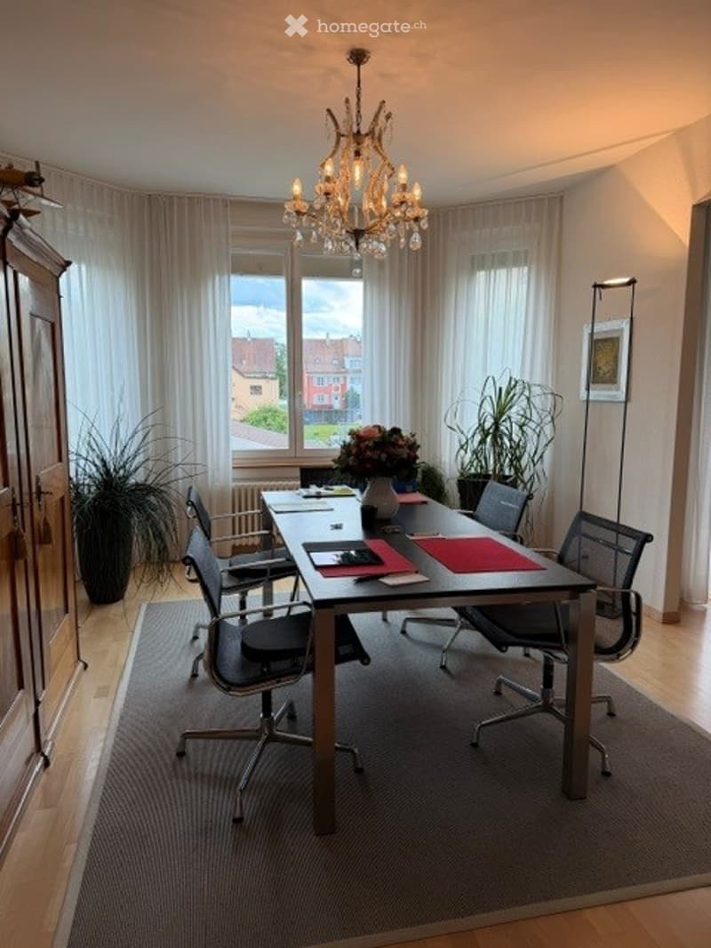 Brightly lit room with large windows offering a view, featuring a central conference table surrounded by chairs, a chandelier hanging above, and decorative plants in the corners.