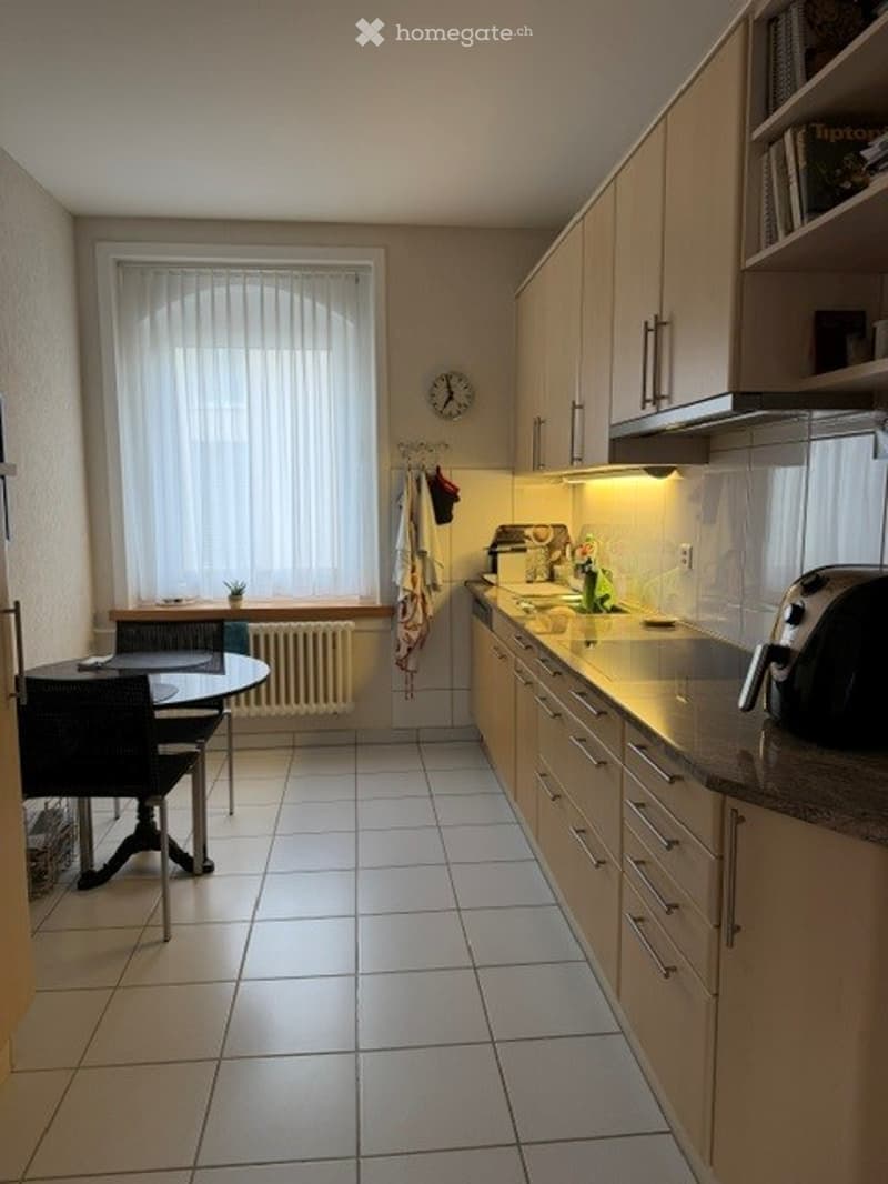 Bright kitchen with white tiled flooring, light wood cabinets, a central island with sink and faucet, a toaster on the counter, a small dining area with a table and chairs, and a window with blinds.