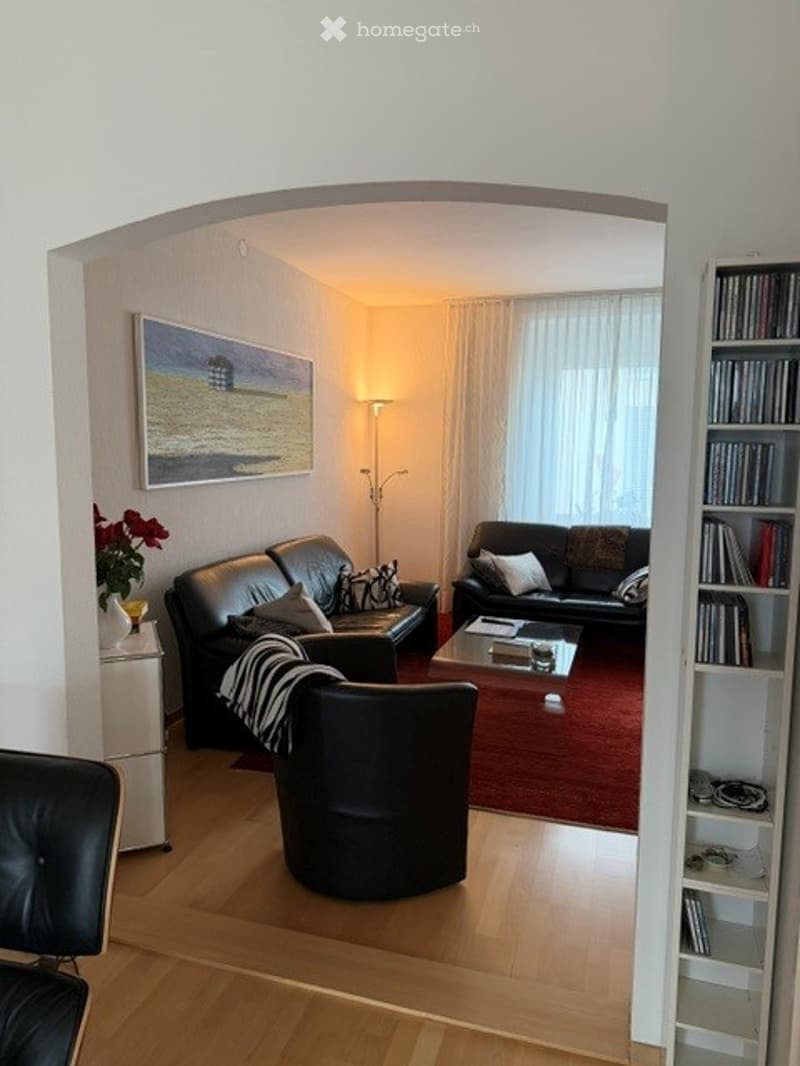 Modern living room with dark leather sofas, a coffee table, a floor lamp, and a bookshelf filled with books. The room features a large window with white curtains and a red area rug.