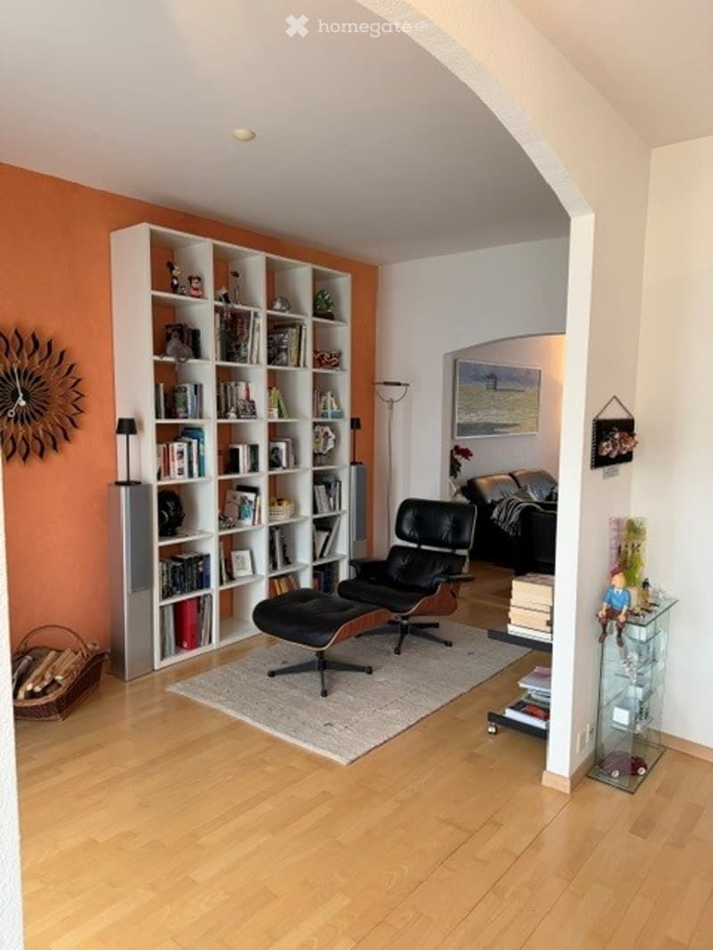 Living room with wooden flooring, white bookshelf filled with books, black leather chair and ottoman, sofa, floor lamp, and decorative items.