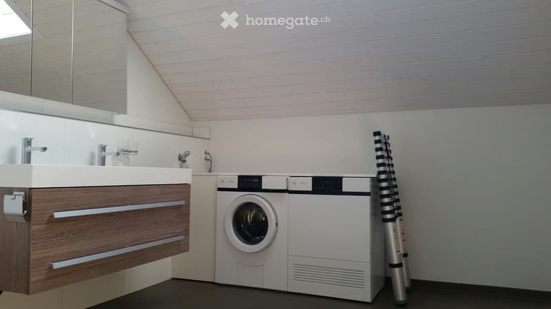 Modern bathroom with a sleek wooden vanity, integrated sink, and a compact washer/dryer unit. A large mirror and a tall, striped stool are also visible.