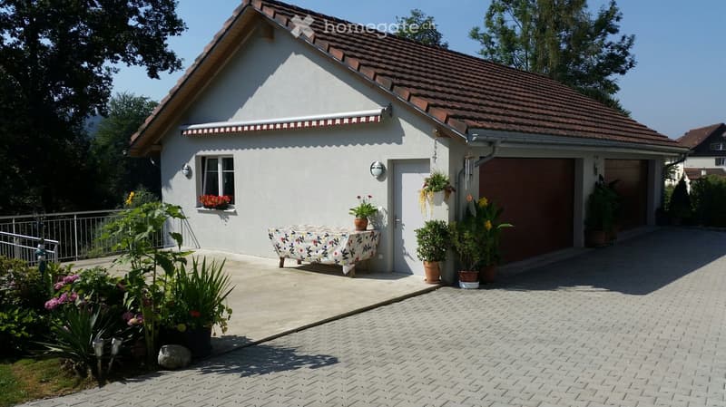 Single-story house with a red-tiled roof, white exterior, garage, and small patio area with potted plants and a table.