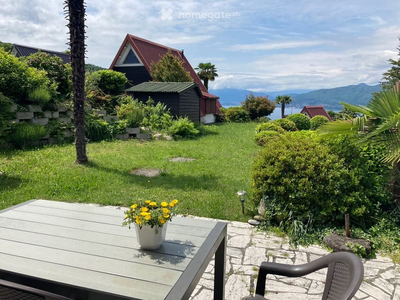 A well-maintained garden with a wooden table, a chair, and a flower pot, surrounded by greenery and a scenic mountain view.