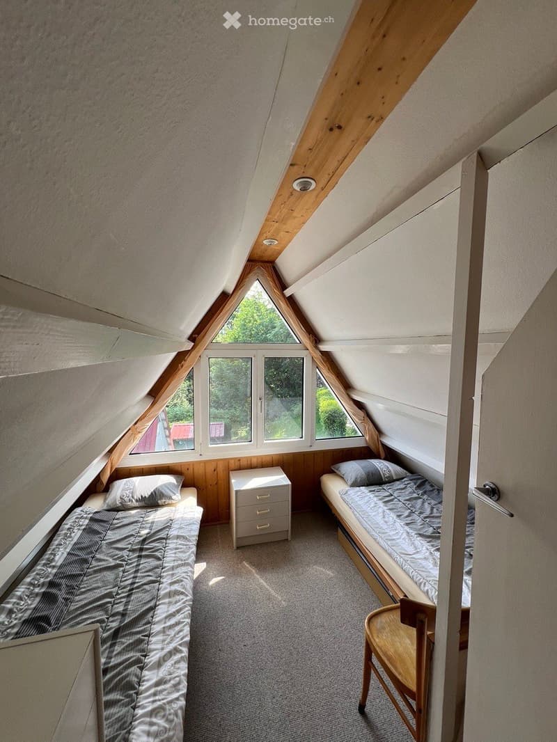 Cozy attic bedroom with two single beds, a nightstand, and a wooden chair, featuring large windows and wooden beams.