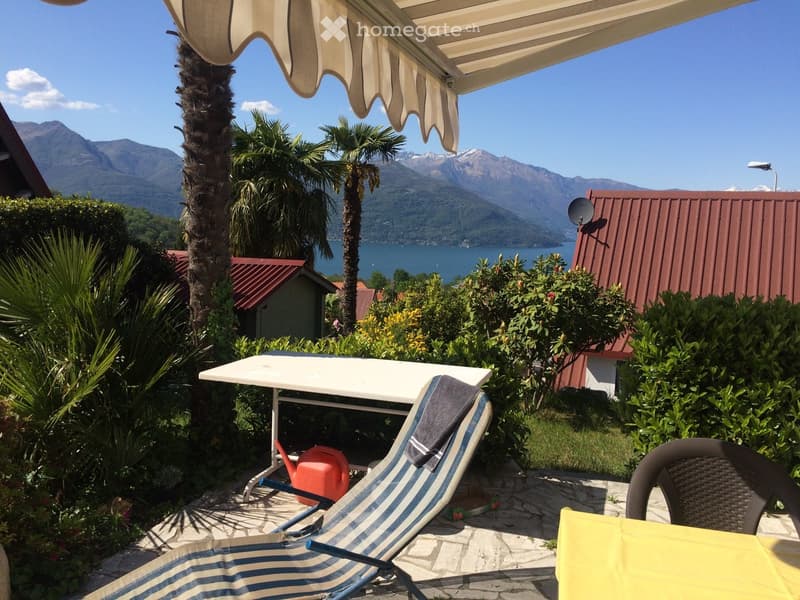 A sunny outdoor patio with a table, chairs, and a lounge chair, overlooking a scenic lake and mountains.
