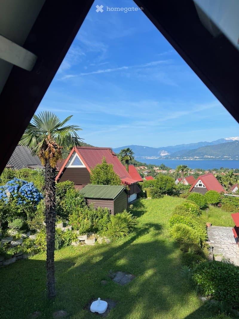 A well-maintained garden with green grass, a palm tree, and several small wooden structures, with a scenic view of a lake and mountains in the background.