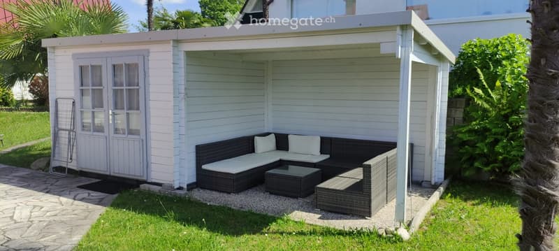 A small, white wooden outdoor structure with a covered seating area featuring a black wicker sofa, white cushions, and a black coffee table.