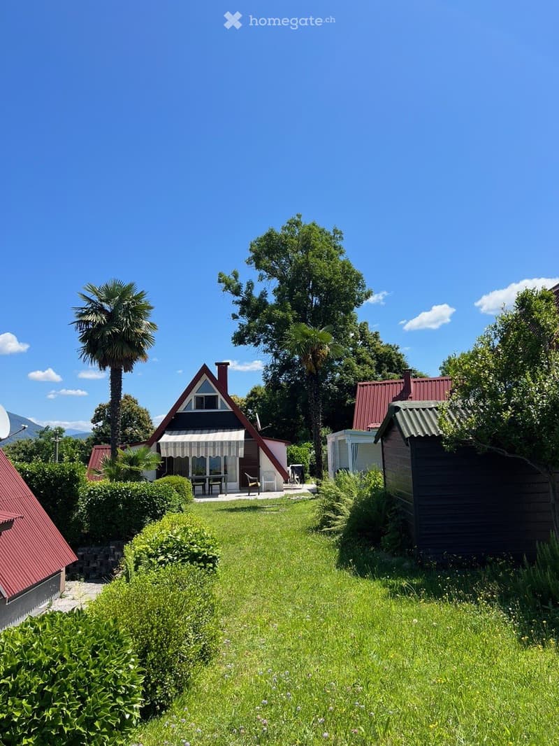 Well-maintained garden with a lawn, trimmed hedges, and several houses with distinct architectural styles, including a triangular roof house and a red-roofed building.