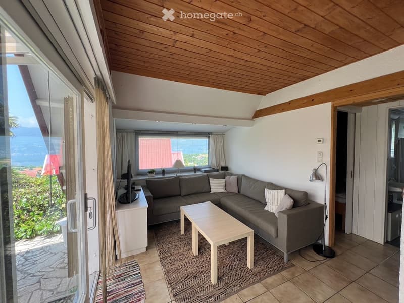 Modern living room with a grey sectional sofa, white coffee table, floor lamp, and television. Large windows offer a scenic view, and the space features a wooden ceiling and tiled floor.