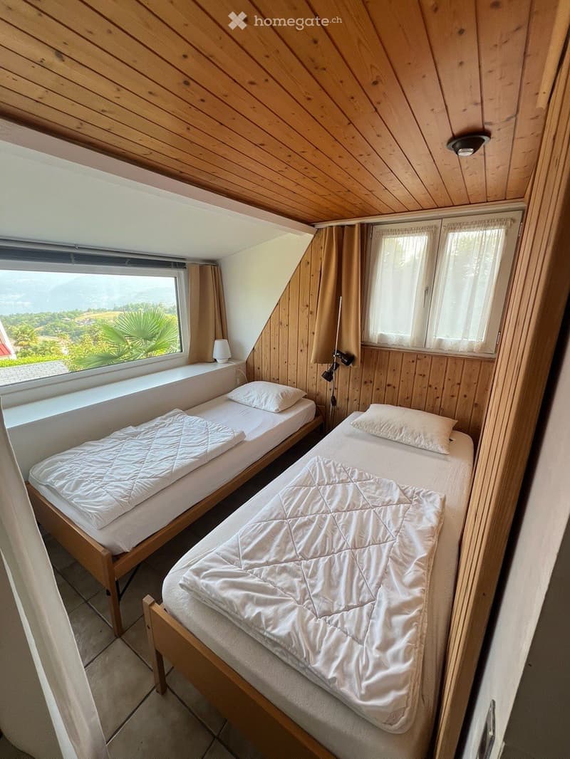Cozy attic bedroom with two single beds, wooden walls, slanted ceiling, and scenic view through large windows.