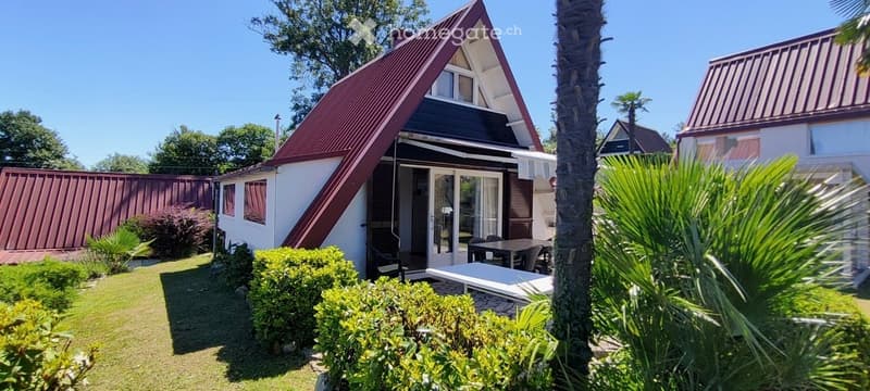 A modern, single-story house with a red metal roof and white walls, surrounded by a well-maintained garden with green lawns, shrubs, and palm trees. The house features large glass doors leading to a patio area with outdoor furniture.