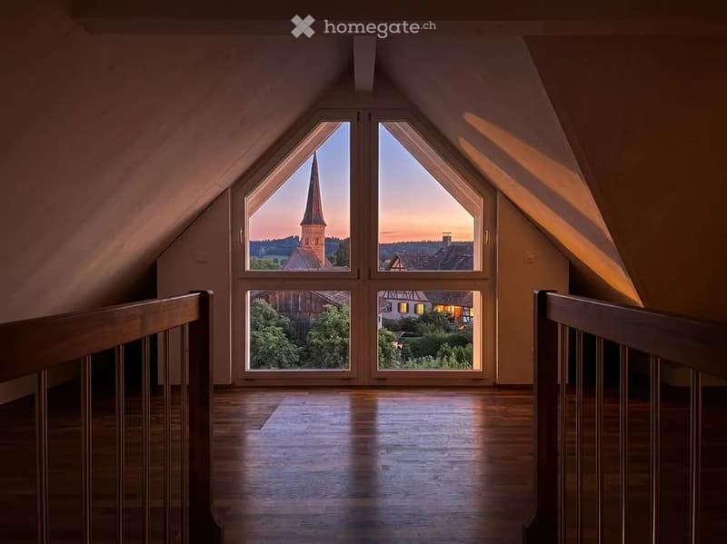 Bright, empty attic space with wooden floors, large windows offering a scenic view of a town and countryside.