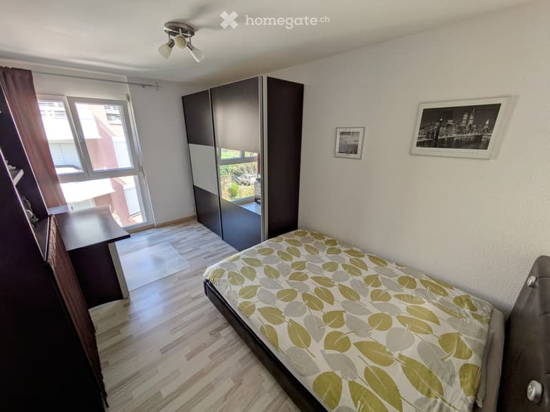 A modern bedroom with a single bed, a large wardrobe with mirrored doors, a table, and framed pictures on the wall. The room features wooden flooring and large windows with curtains.