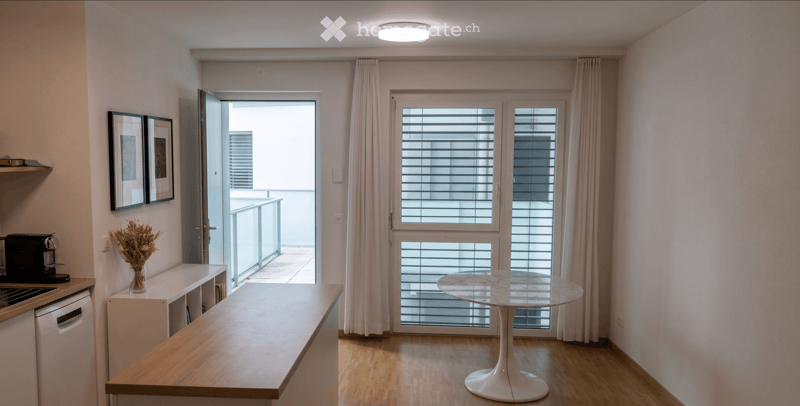 Modern kitchen and dining area with a round white table, coffee maker, dishwasher, and a balcony with blinds.