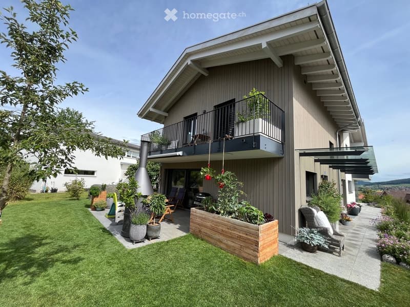 Well-maintained garden with a lawn, wooden planter box, outdoor seating, and a modern house with balconies and a chimney.