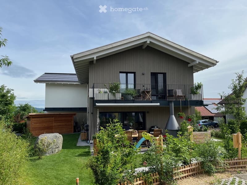 Modern two-story house with grey siding, large windows, balcony, and well-maintained garden with patio furniture and barbecue.
