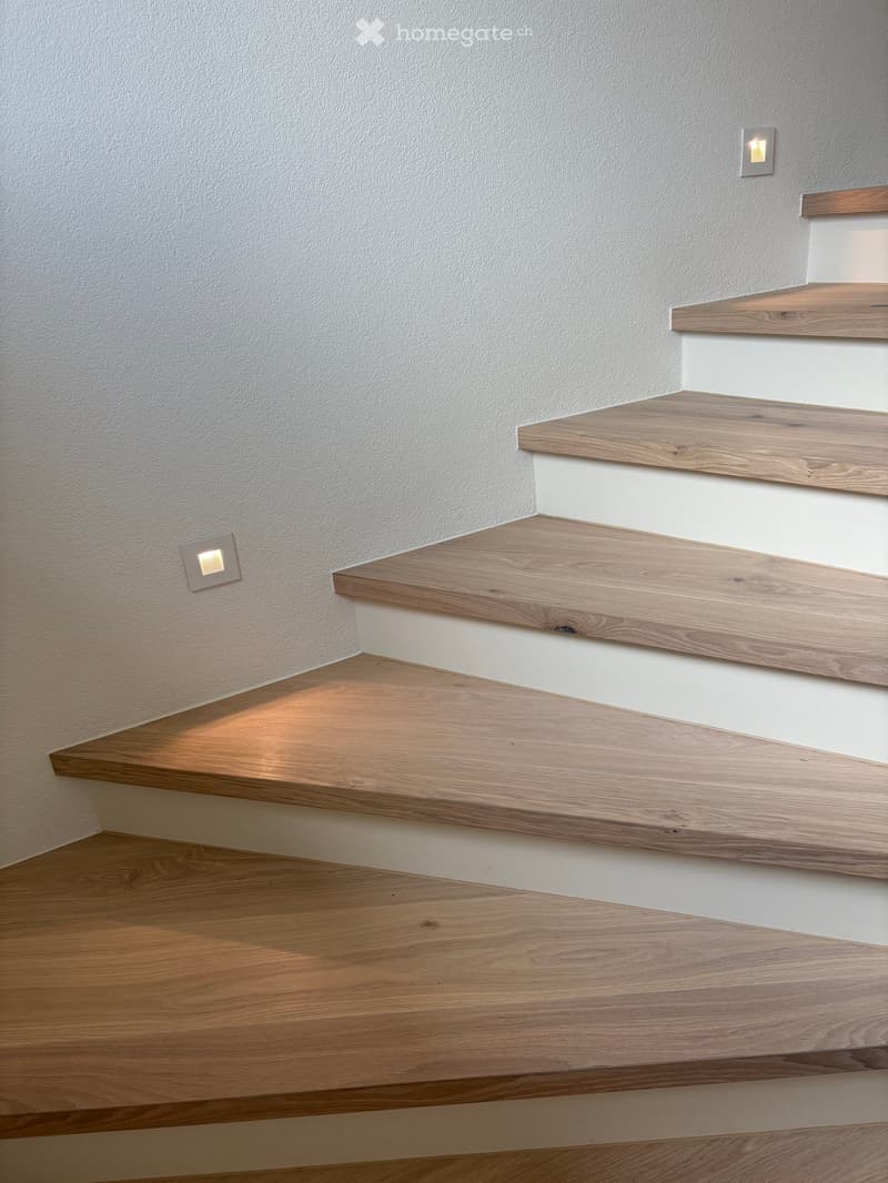 Modern staircase with wooden steps and white risers, illuminated by wall-mounted lights.