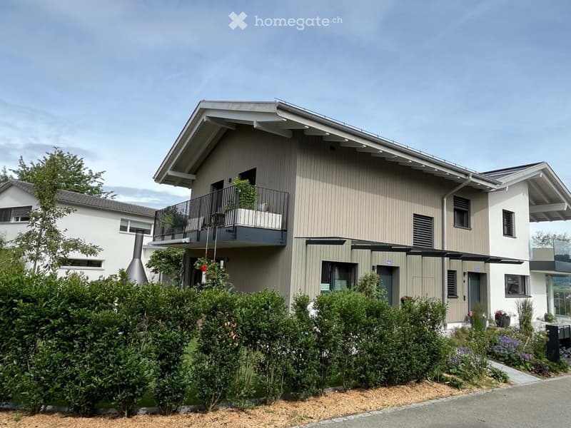 Modern two-story house with beige siding, large windows, and a balcony on the upper level. The house features a well-maintained garden with shrubs and flowers in front.