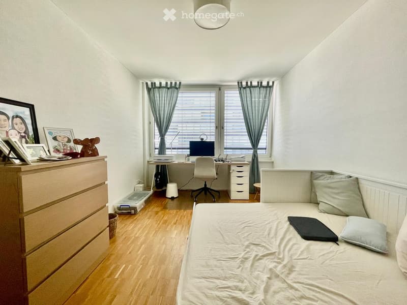 A cozy bedroom with a bed, desk, and chair. The room features a wooden floor, white walls, and large windows with curtains. A dresser with picture frames and a small stool are also visible.