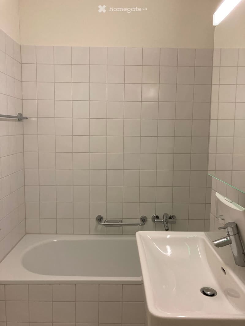 Bathroom with a bathtub, sink, and handrail. The walls are tiled in light gray, and the fixtures are modern and silver.