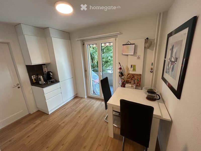 A small, modern kitchen area with white cabinets, a small dining table with chairs, and a coffee maker on the counter. There is a sliding glass door leading to a balcony.