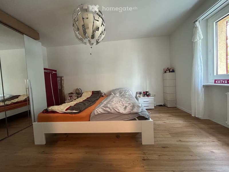 Bedroom with wooden flooring, a white bed with orange and grey bedding, a large mirror wardrobe, a nightstand with a lamp, and a window with white curtains.