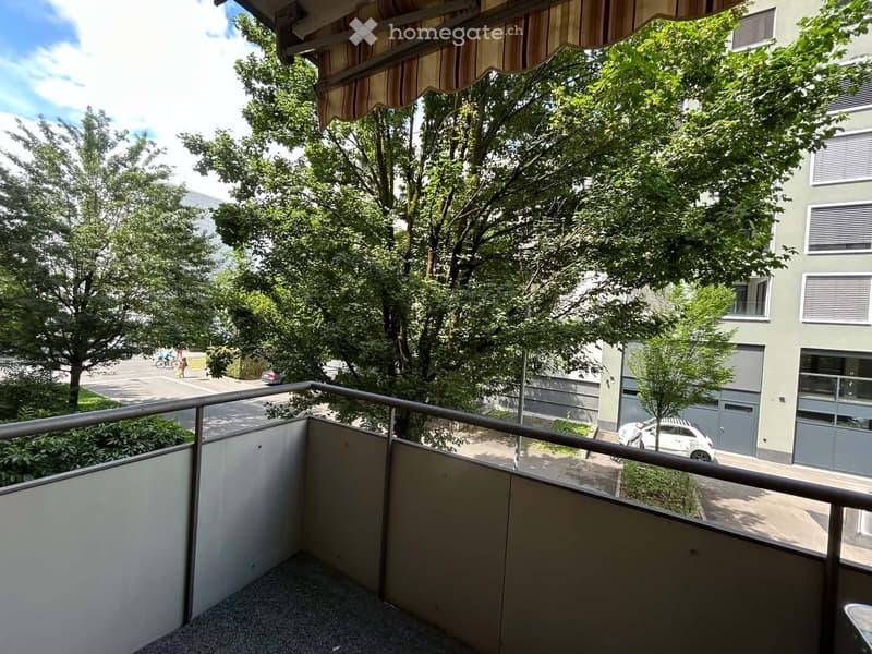 Balcony with metal railing, striped awning, overlooking a tree-lined street with parked cars and buildings.