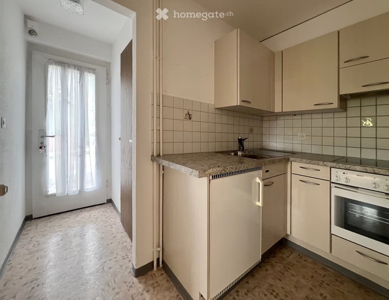 Modern kitchen with light-colored cabinets, granite countertop, sink, and built-in oven. Adjacent hallway with tiled floor and window with white curtains.