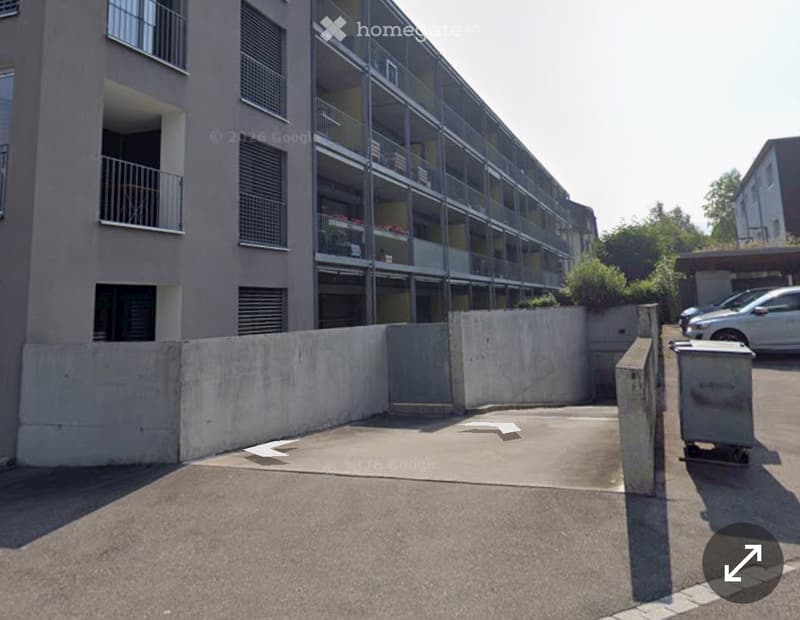 Modern multi-story building with large glass windows and balconies, surrounded by a concrete wall and parking area.