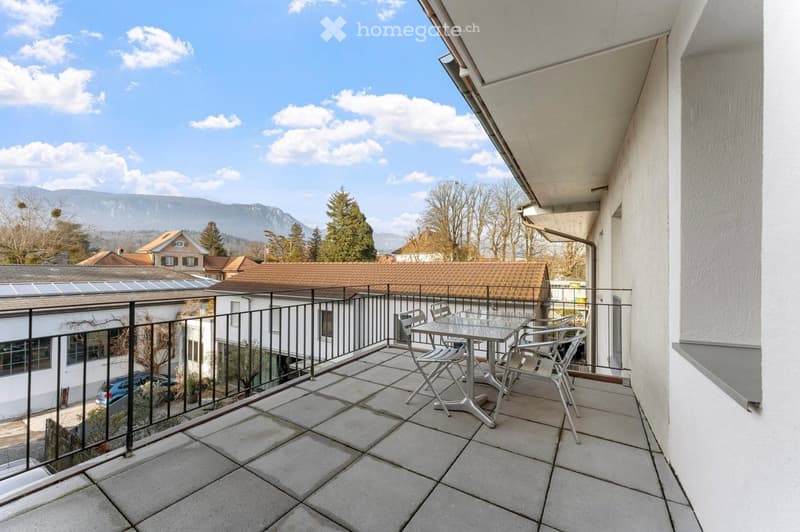 Balcony with tiled floor, metal railing, and a small table with chairs, overlooking a scenic view of mountains and houses.