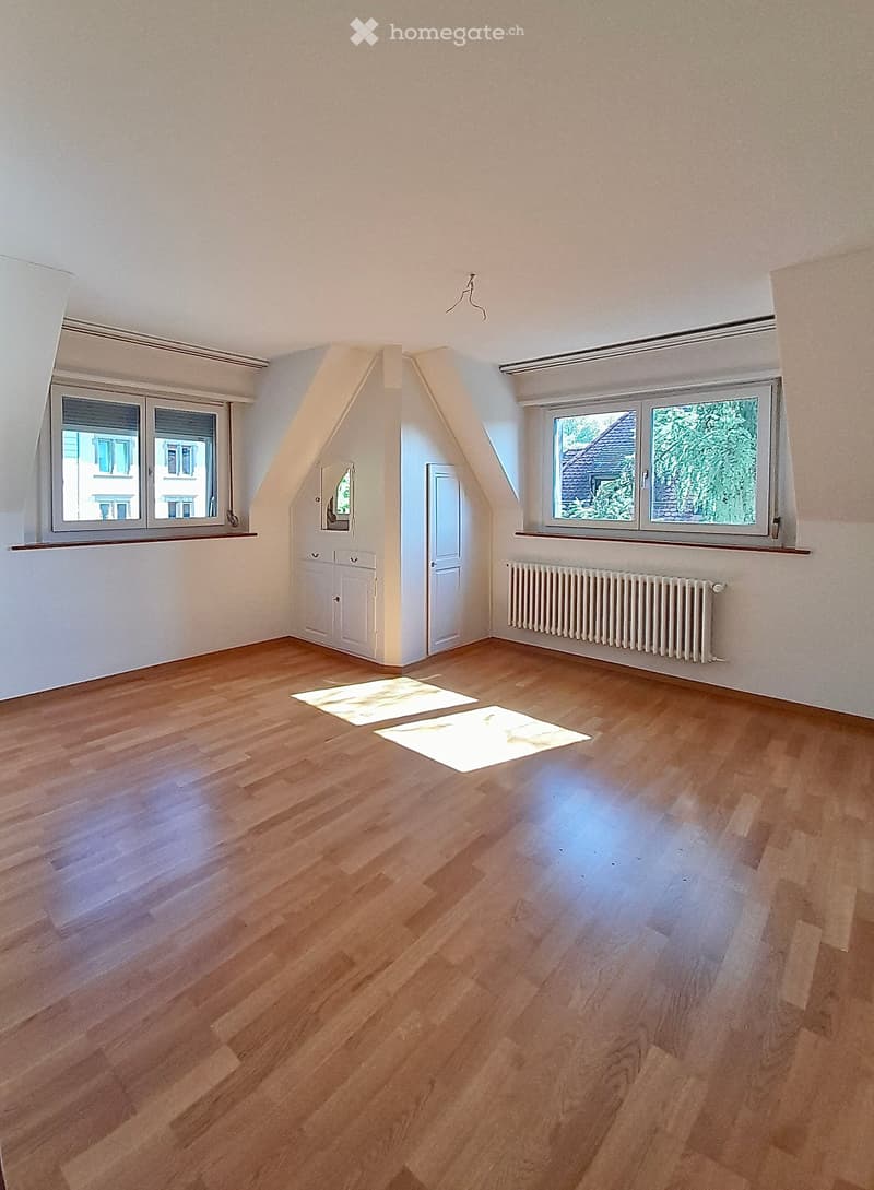 Empty attic room with wooden flooring, white walls, two windows, a door, and a radiator. The room has a slanted ceiling typical of attic spaces.