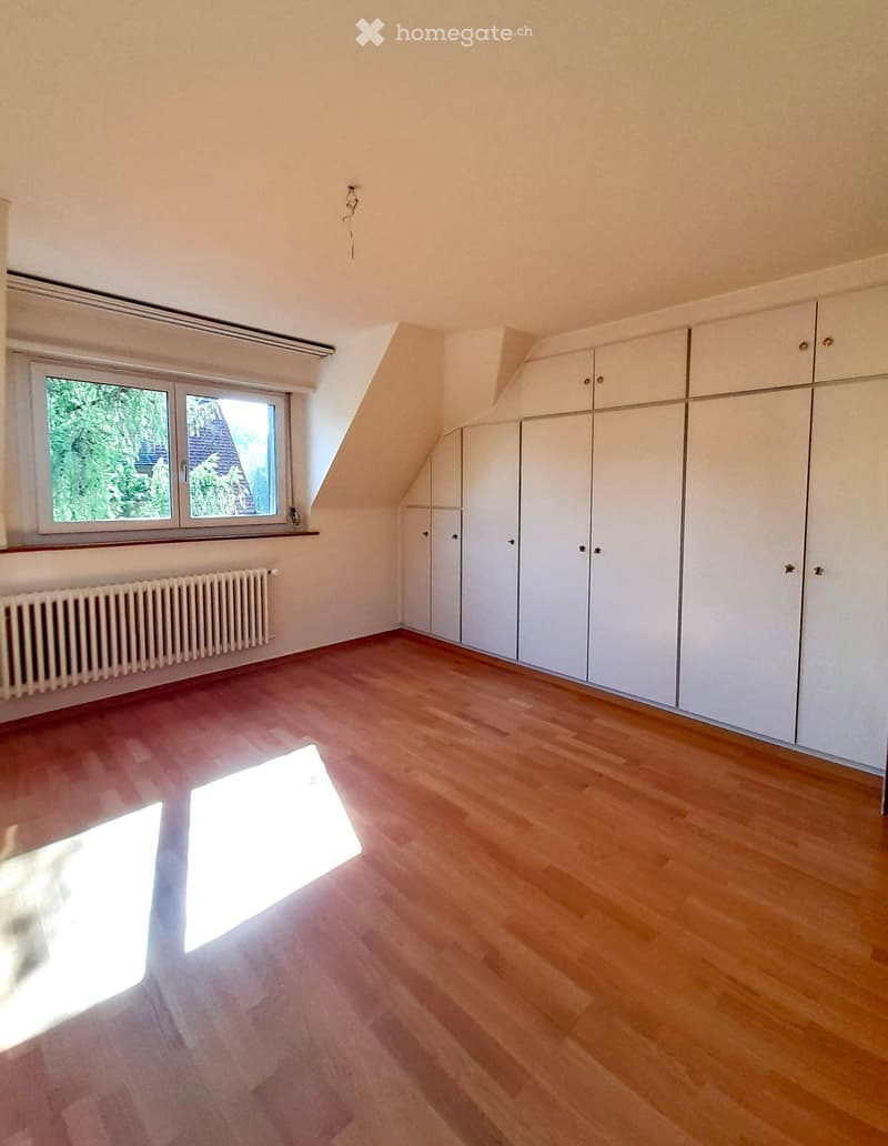 Empty attic room with wooden flooring, white built-in wardrobes, and a window offering a view of greenery outside.