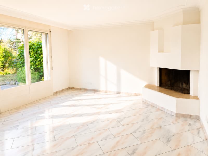 Empty room with white walls, marble tiled floor, large windows with greenery view, and a built-in fireplace.