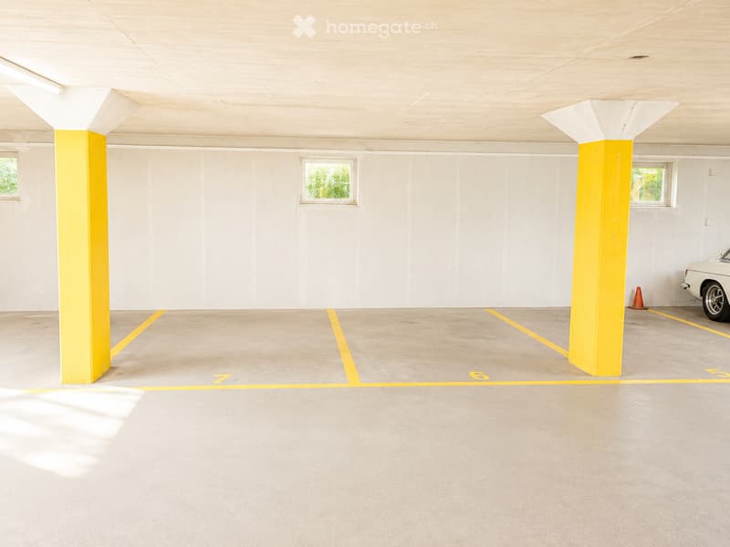 Underground parking area with concrete floors, white walls, yellow pillars, and numbered parking spaces. A white vintage car is partially visible in one of the parking spots.